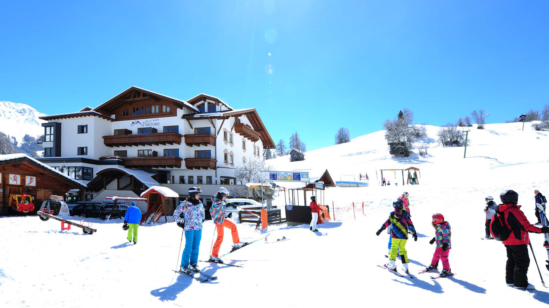 Skiende Gäste und Kinder vor dem 4-Sterne-Hotel Panorama in verschneiter Berglandschaft unter klarem Himmel.